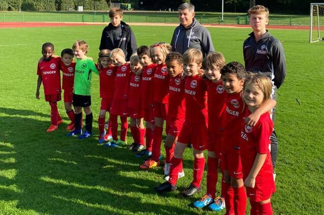 Gruppe von Kindern in roten Fu&szlig;balltrikots mit Trainern auf einem Sportplatz, umgeben von B&auml;umen und einem Tor.