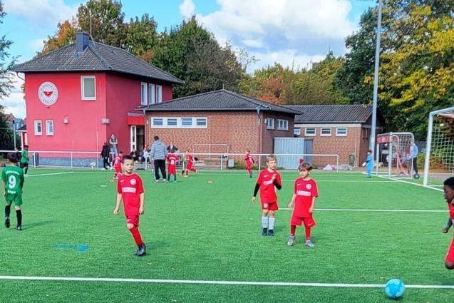 Sechs Kinder in roten und gr&uuml;nen Trikots spielen Fu&szlig;ball auf einem Kunstrasenplatz vor einem Vereinsgeb&auml;ude.