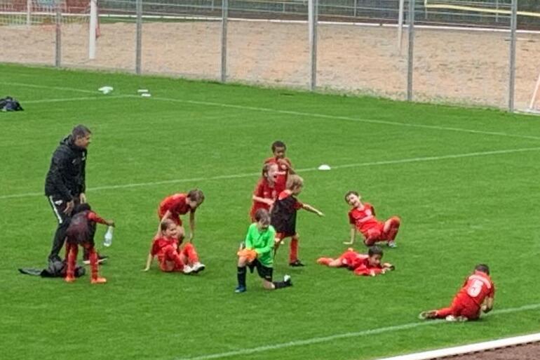 Eine Gruppe von Kindern in roten Trikots trainiert auf einem Fu&szlig;ballplatz, w&auml;hrend ein Trainer Anweisungen gibt.