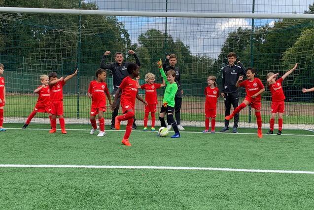 Gruppe von Kindern in roten Fu&szlig;balltrikots steht vor einem Tor; ein Torwart bereitet sich auf einen Schuss vor.