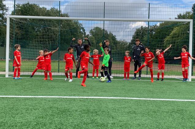 Gruppe von Kindern in roten Fu&szlig;balltrikots steht vor einem Tor; ein Torwart bereitet sich auf einen Schuss vor.