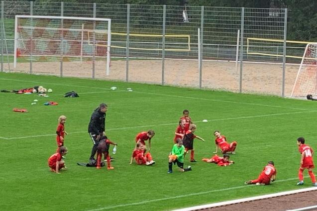 Eine Gruppe von Kindern in roten Trikots trainiert auf einem Fu&szlig;ballplatz, w&auml;hrend ein Trainer Anweisungen gibt.