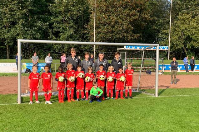 Gruppe von Fu&szlig;ballspielern in roten Trikots steht vor einem Tor, umgeben von einem Sportplatz und Zuschauern.