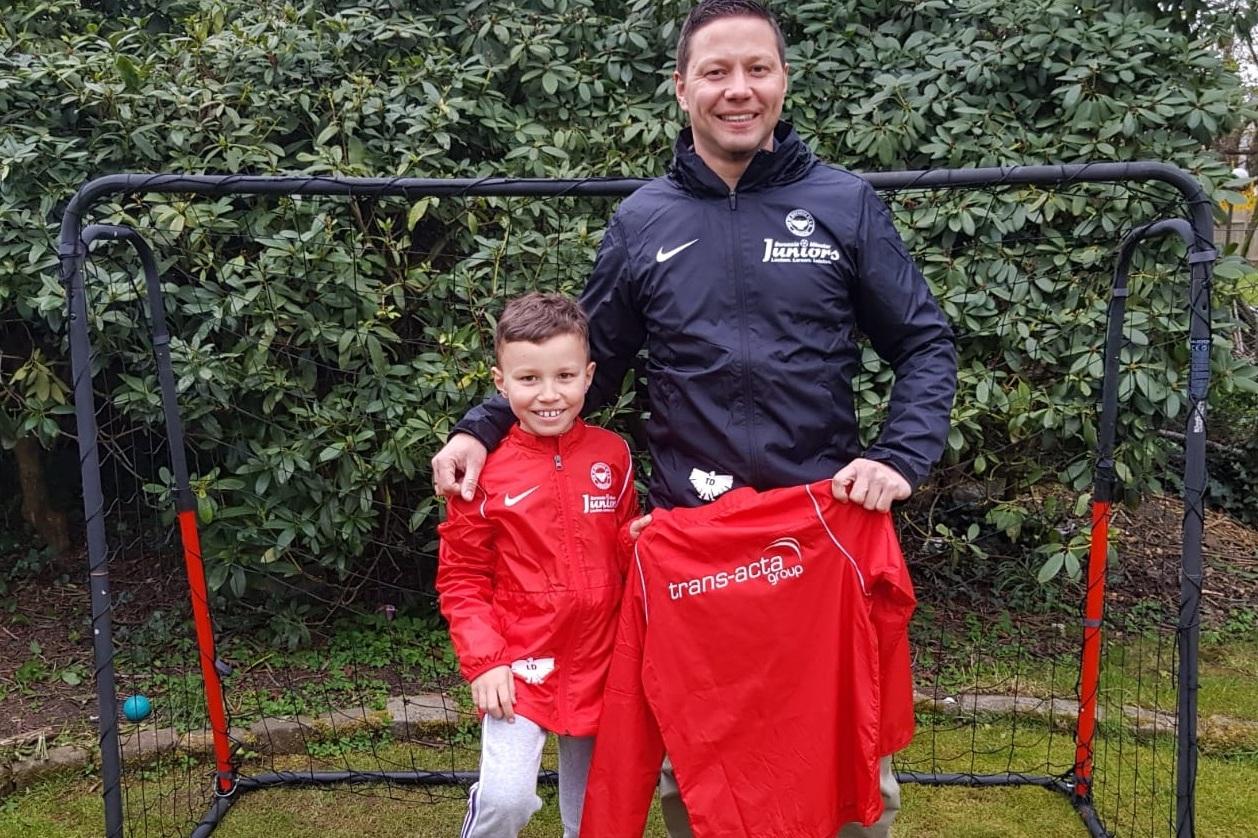 Vater und Sohn stehen vor einem Fu&szlig;balltor, beide in roten Trainingsjacken, der Sohn h&auml;lt ein Fu&szlig;balltrikot in der Hand.
