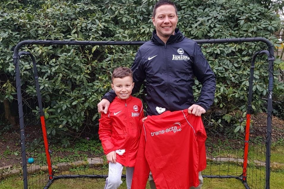 Vater und Sohn stehen vor einem Fu&szlig;balltor, beide in roten Trainingsjacken, der Sohn h&auml;lt ein Fu&szlig;balltrikot in der Hand.