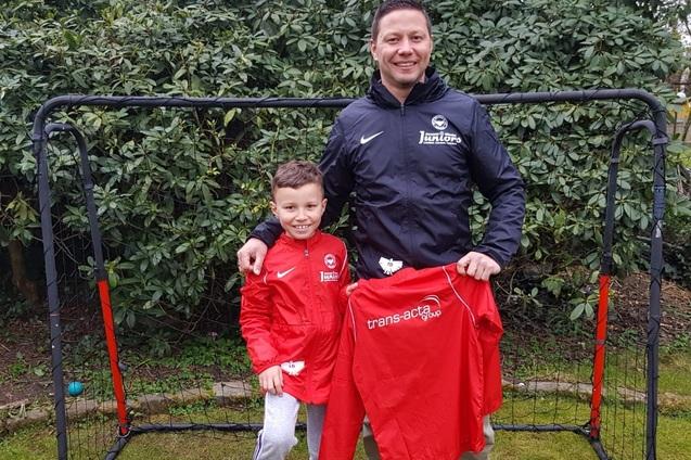 Vater und Sohn stehen vor einem Fu&szlig;balltor, beide in roten Trainingsjacken, der Sohn h&auml;lt ein Fu&szlig;balltrikot in der Hand.