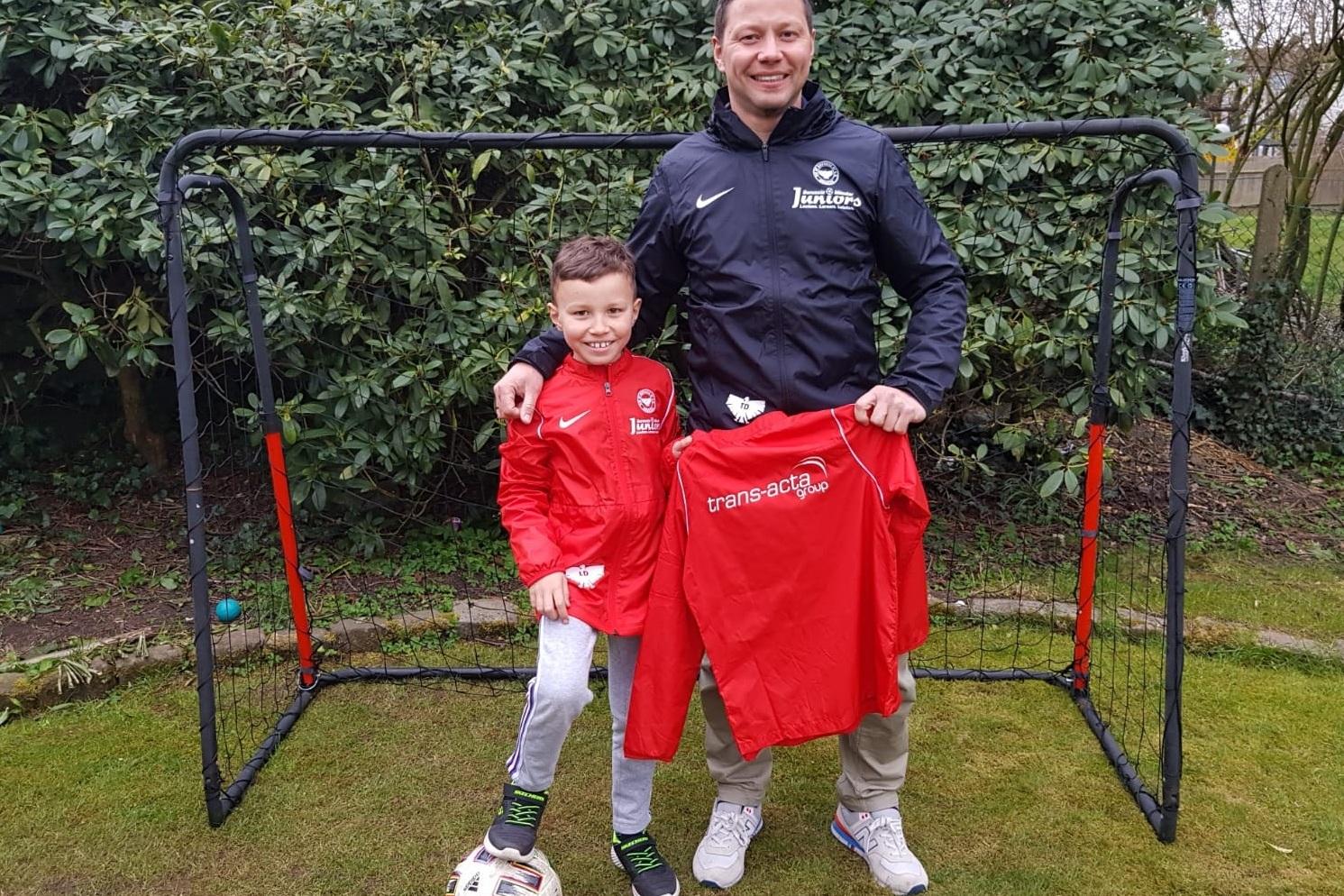 Vater und Sohn stehen vor einem Fu&szlig;balltor, beide in roten Trainingsjacken, der Sohn h&auml;lt ein Fu&szlig;balltrikot in der Hand.