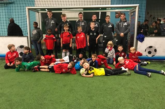 Gruppenfoto von Jungen in Sportkleidung vor einem Fu&szlig;balltor in einer Halle, einige liegen auf dem Rasen, andere stehen.