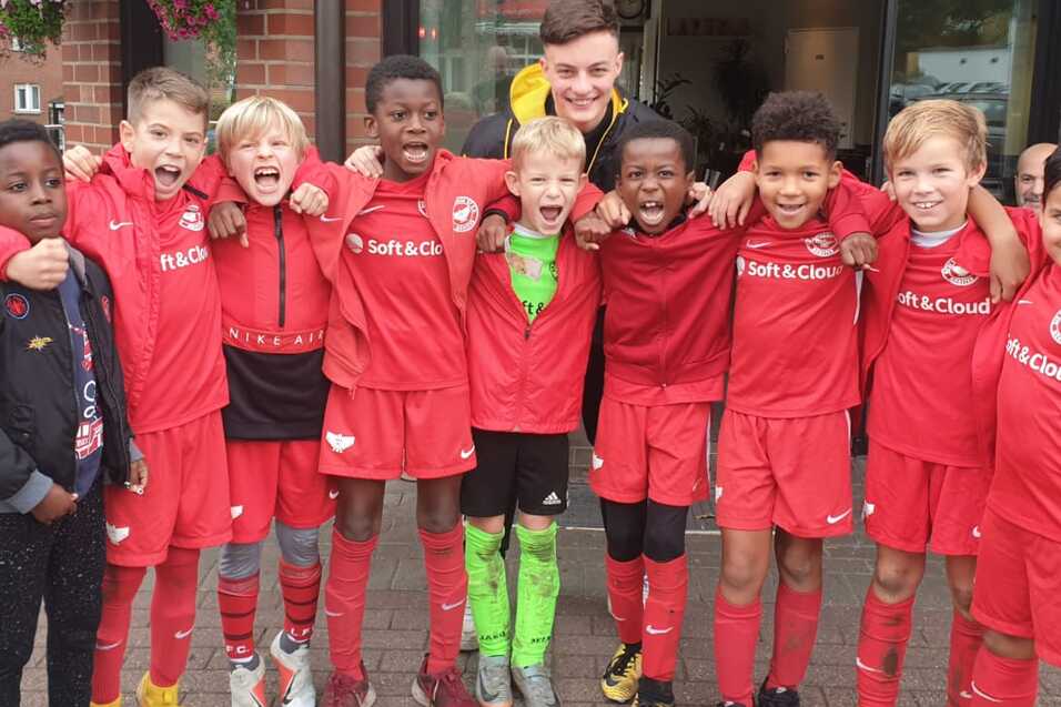 Gruppe von zehn jungen Fu&szlig;ballspielern in roten Trikots posiert fr&ouml;hlich vor einem Vereinsgeb&auml;ude.