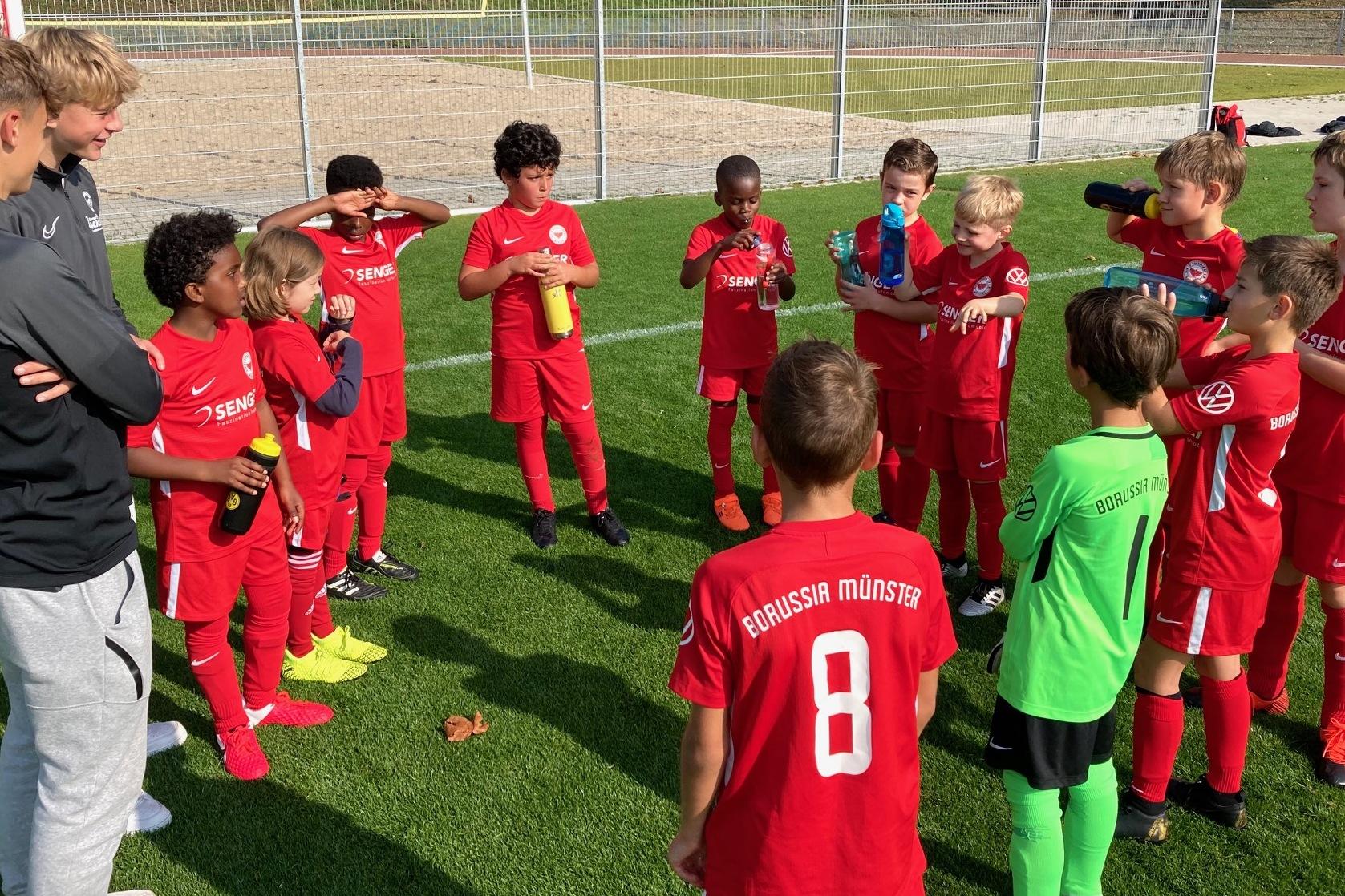 Gruppe von jungen Fu&szlig;ballspielern in roten Trikots versammelt sich um einen Trainer auf einem Sportplatz.