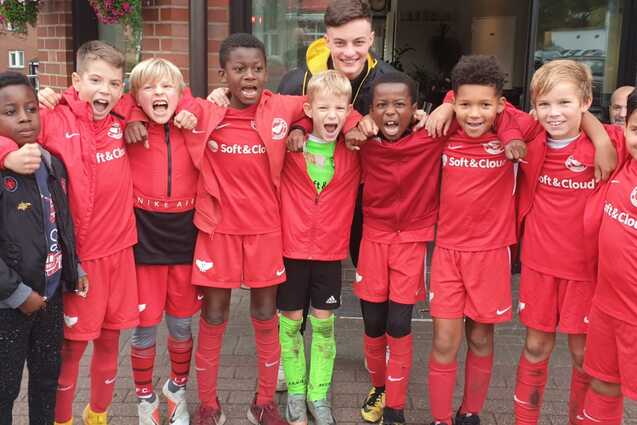 Gruppe von zehn jungen Fu&szlig;ballspielern in roten Trikots posiert fr&ouml;hlich vor einem Vereinsgeb&auml;ude.