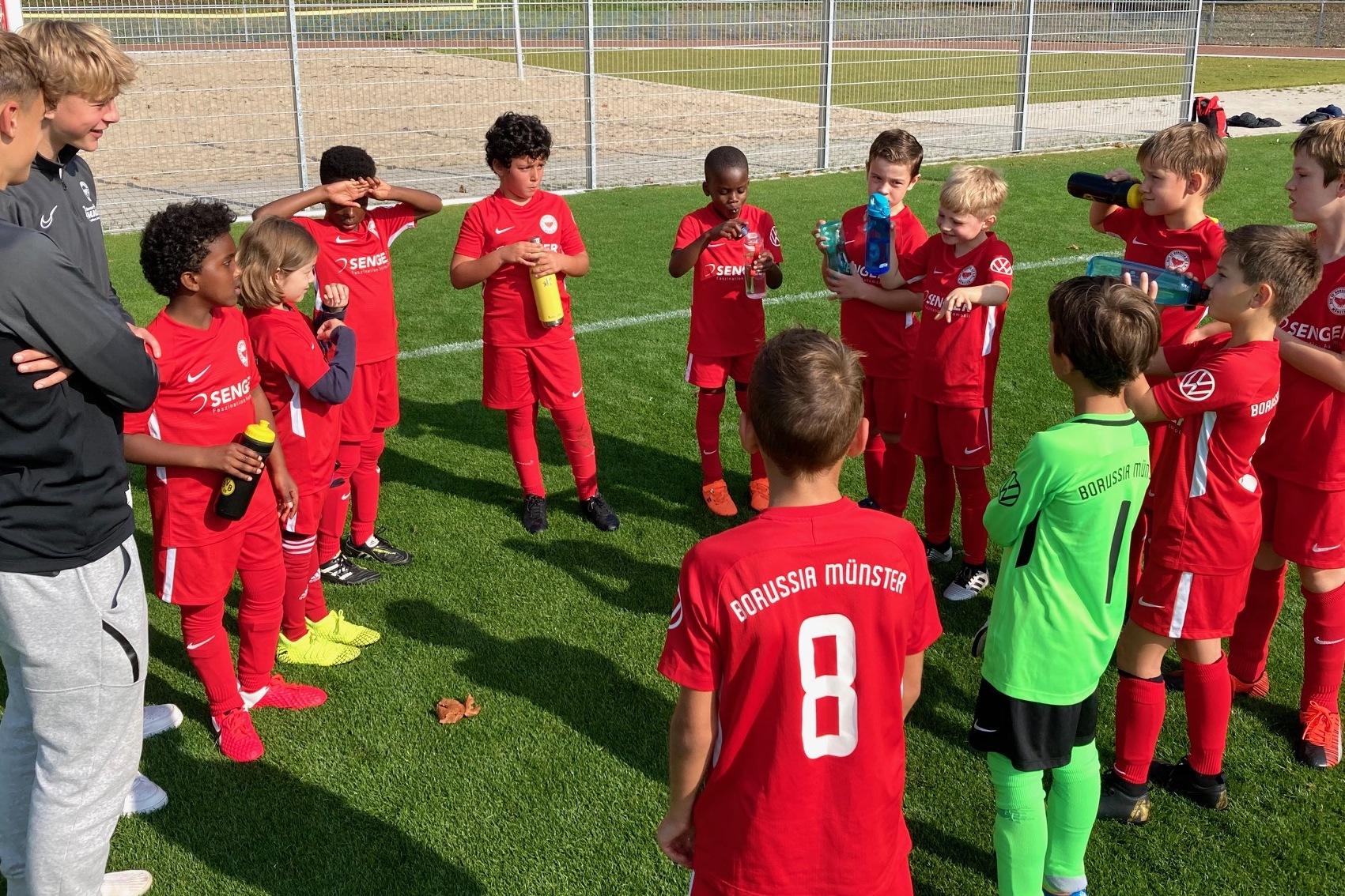 Gruppe von jungen Fu&szlig;ballspielern in roten Trikots versammelt sich um einen Trainer auf einem Sportplatz.