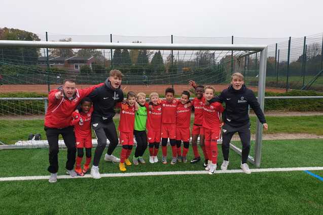 Gruppe junger Fu&szlig;ballspieler in roten Trikots steht l&auml;chelnd vor einem Tor auf einem Fu&szlig;ballplatz.