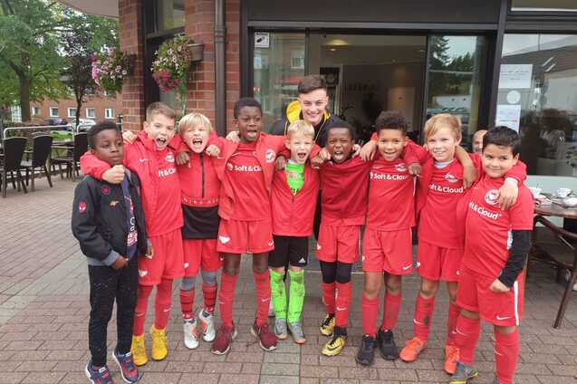 Gruppe von zehn jungen Fu&szlig;ballspielern in roten Trikots posiert fr&ouml;hlich vor einem Vereinsgeb&auml;ude.