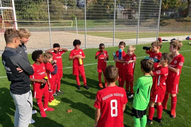 Gruppe von jungen Fu&szlig;ballspielern in roten Trikots versammelt sich um einen Trainer auf einem Sportplatz.