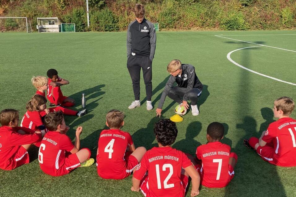 Zehn Kinder in roten Fu&szlig;balltrikots sitzen um einen Trainer auf einem gr&uuml;nen Fu&szlig;ballfeld und h&ouml;ren aufmerksam zu.