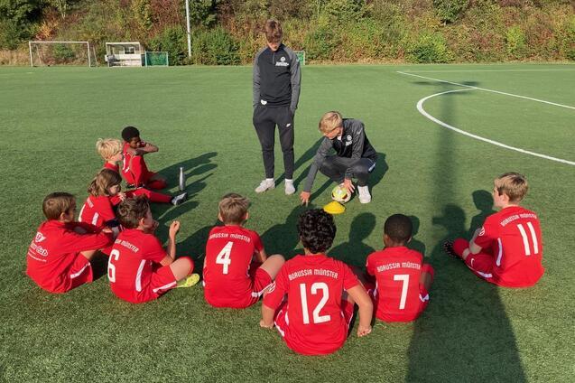 Zehn Kinder in roten Fu&szlig;balltrikots sitzen um einen Trainer auf einem gr&uuml;nen Fu&szlig;ballfeld und h&ouml;ren aufmerksam zu.
