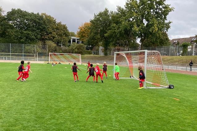 Gruppe von Kindern in roten Trikots beim Fu&szlig;ballspiel vor zwei Toren auf einem Rasenplatz.