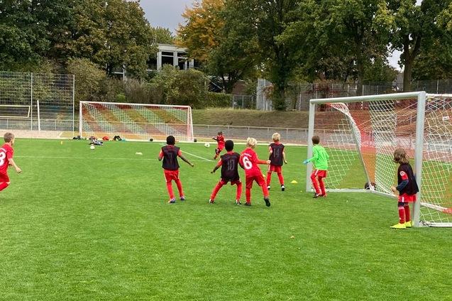 Gruppe von Kindern in roten Trikots beim Fu&szlig;ballspiel vor zwei Toren auf einem Rasenplatz.