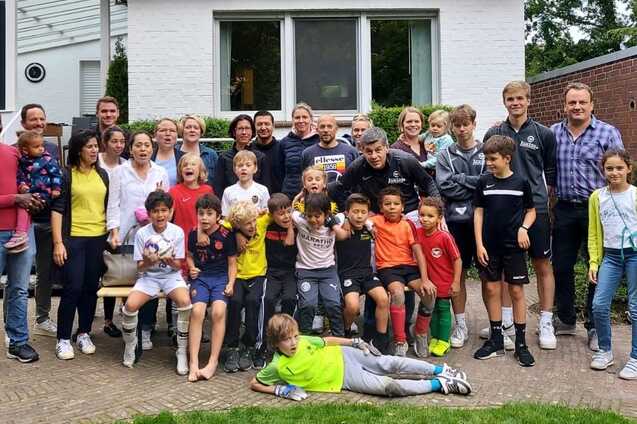 Gruppe von Erwachsenen und Kindern versammelt vor einem Haus, l&auml;chelnd und posierend auf einer Rasenfl&auml;che.