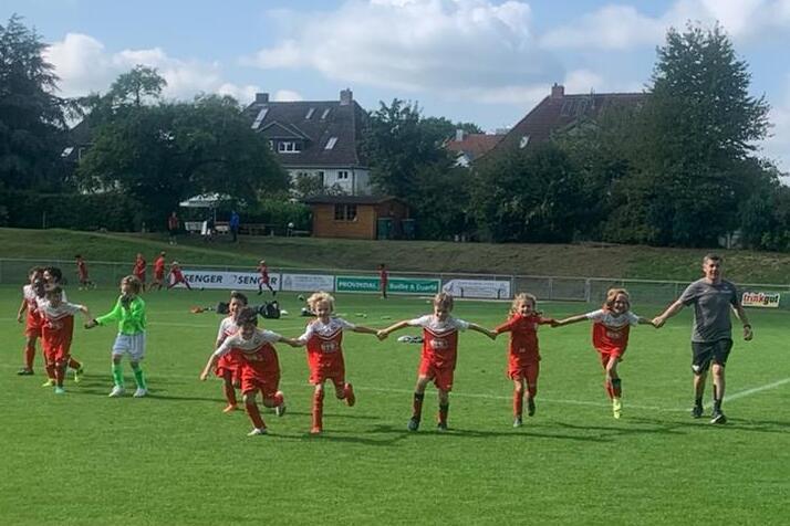Gruppe von Kindern in roten Fu&szlig;balltrikots, die Hand in Hand auf einem gr&uuml;nen Spielfeld laufen, begleitet von einem Trainer.