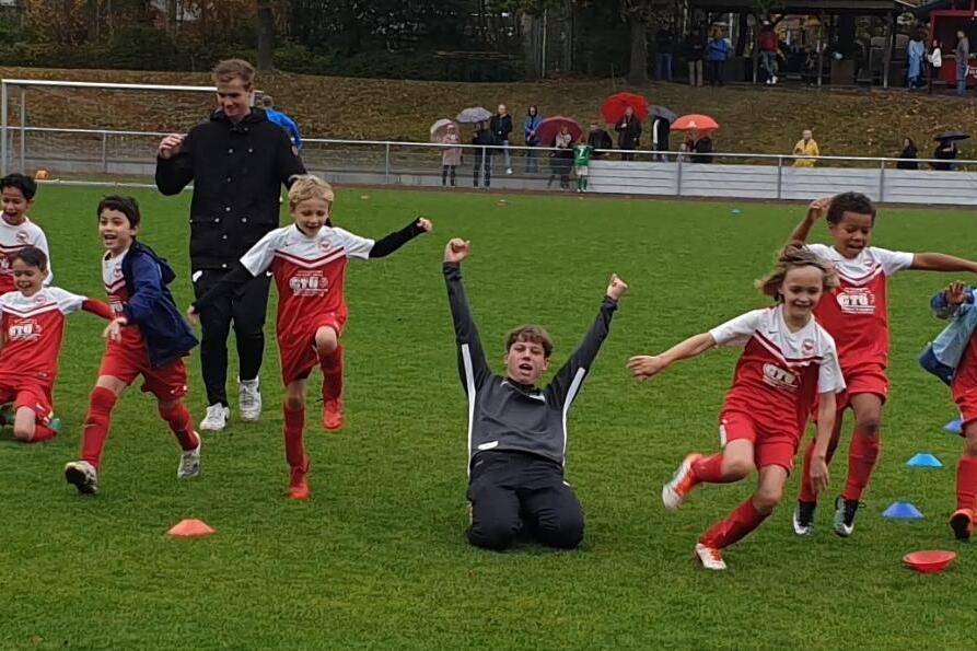 Gruppenszene von jubelnden Kindern in roten Fu&szlig;balltrikots auf einem Rasenplatz, einige Spieler liegen und anderen feiern.