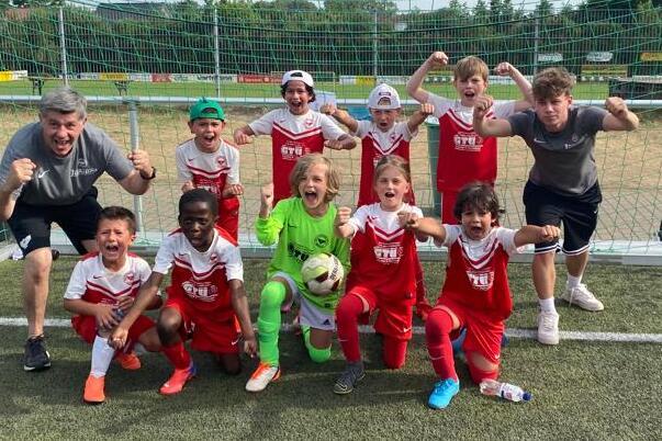 Gruppe von Kindern in roten Fu&szlig;balltrikots, lachend und jubelnd vor einem Tor auf einem Kunstrasenfeld.