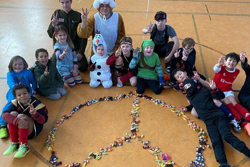 Gruppe von Kindern und Jugendlichen posiert um ein aus S&uuml;&szlig;igkeiten gelegtens Friedenssymbol in einer Sporthalle.