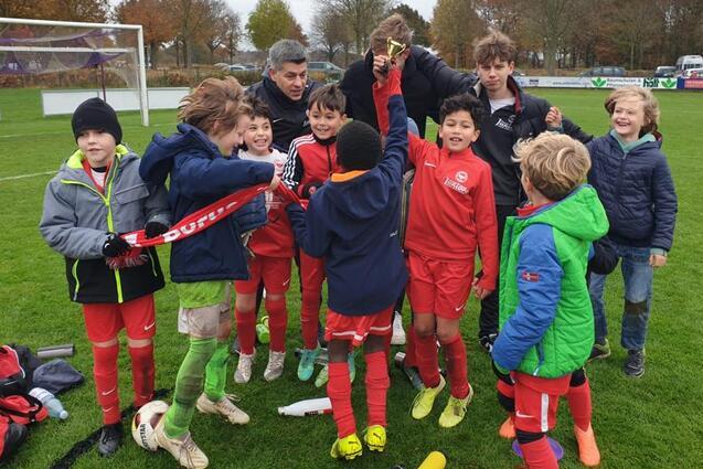Gruppe von Kindern in Fu&szlig;balloutfits, die gemeinsam einen Pokal auf einem Sportplatz feiern.