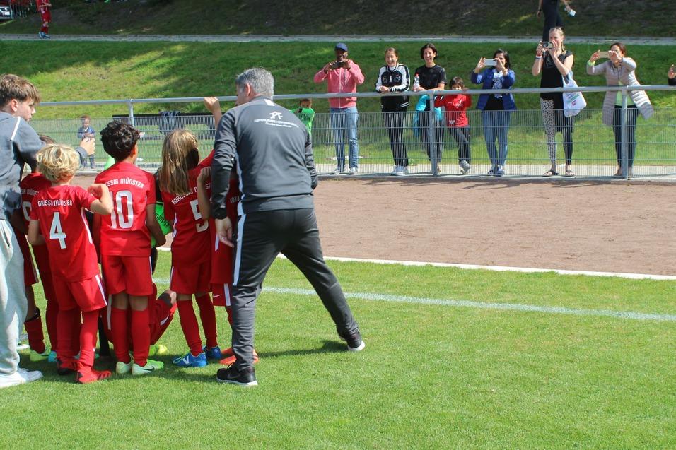 Gruppierung von Kindern in roten Fu&szlig;balltrikots mit Trainer auf einem Spielfeld, Zuschauer applaudieren im Hintergrund.