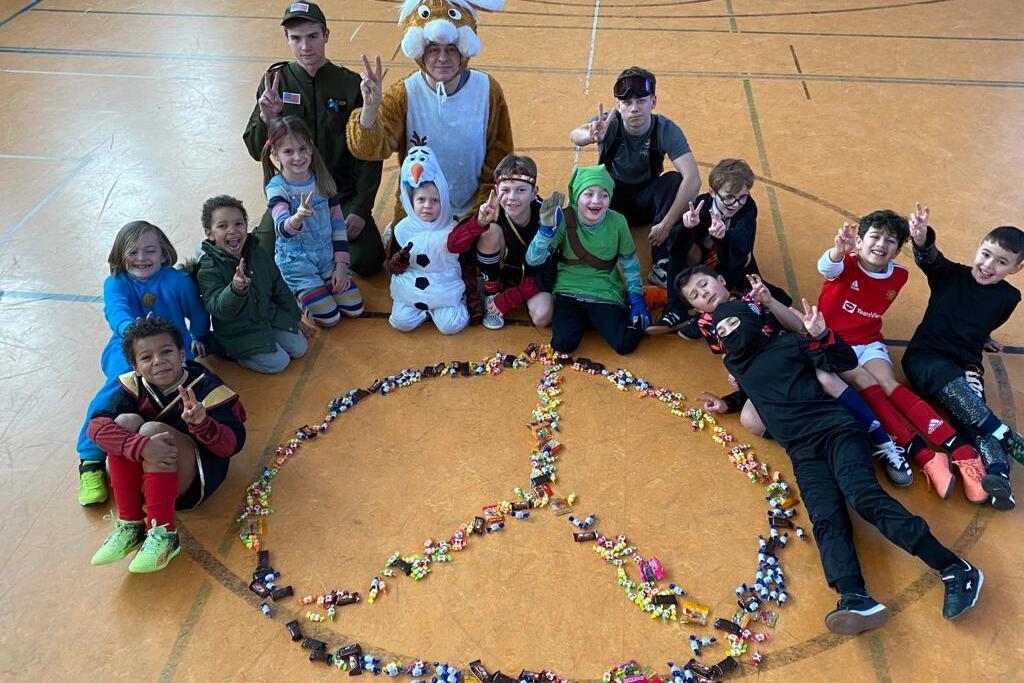 Gruppe von Kindern und Jugendlichen posiert um ein aus S&uuml;&szlig;igkeiten gelegtens Friedenssymbol in einer Sporthalle.