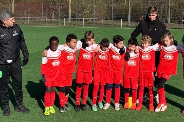 Mannschaft von zehn Fu&szlig;ballkindern in roten Trikots und ein Trainer auf einem Kunstrasenplatz.