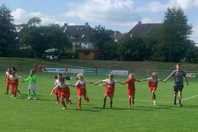 Gruppe von Kindern in roten Fu&szlig;balltrikots, die Hand in Hand auf einem gr&uuml;nen Spielfeld laufen, begleitet von einem Trainer.