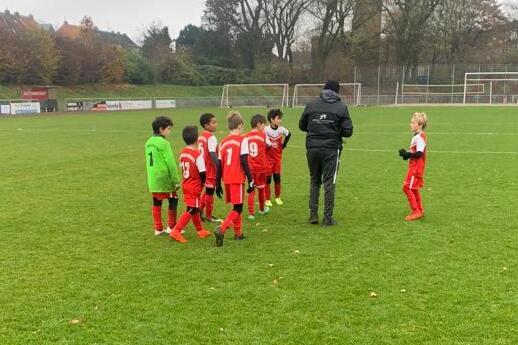Gruppe von Kindern in roten Fu&szlig;balltrikots und ein Trainer auf einem gr&uuml;nen Fu&szlig;ballplatz an einem grauen Tag.
