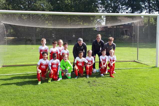 Gruppenfoto einer Jugendfu&szlig;ballmannschaft in roten Trikots, stehend und kniend vor einem Tor auf dem Rasenplatz.