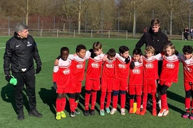 Mannschaft von zehn Fu&szlig;ballkindern in roten Trikots und ein Trainer auf einem Kunstrasenplatz.