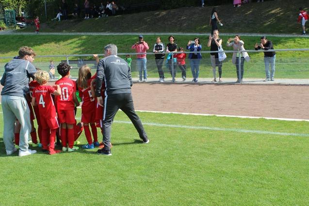 Gruppierung von Kindern in roten Fu&szlig;balltrikots mit Trainer auf einem Spielfeld, Zuschauer applaudieren im Hintergrund.