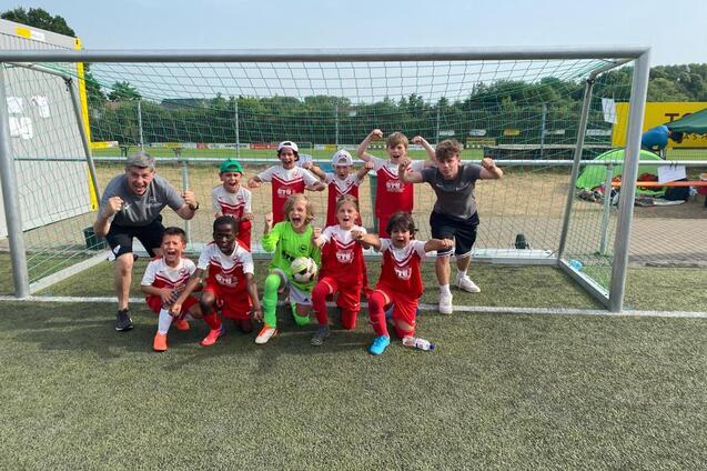 Gruppe von Kindern in roten Fu&szlig;balltrikots, lachend und jubelnd vor einem Tor auf einem Kunstrasenfeld.