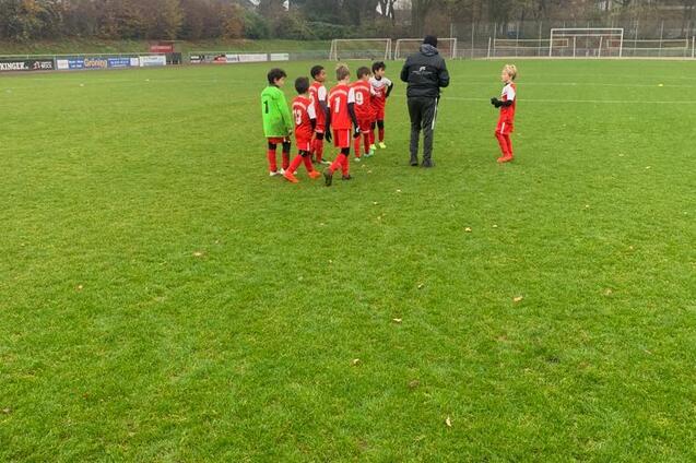Gruppe von Kindern in roten Fu&szlig;balltrikots und ein Trainer auf einem gr&uuml;nen Fu&szlig;ballplatz an einem grauen Tag.