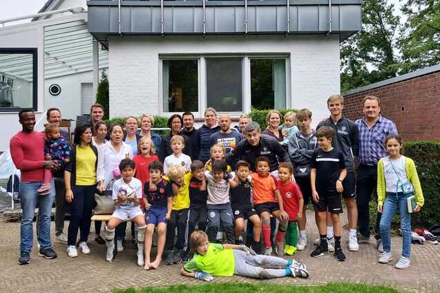 Gruppe von Erwachsenen und Kindern versammelt vor einem Haus, l&auml;chelnd und posierend auf einer Rasenfl&auml;che.