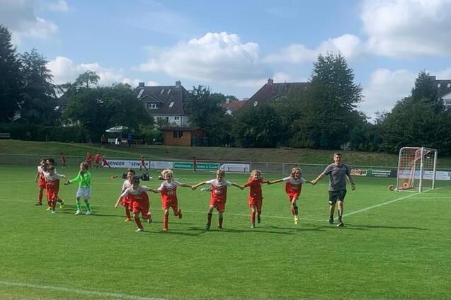 Gruppe von Kindern in roten Fu&szlig;balltrikots, die Hand in Hand auf einem gr&uuml;nen Spielfeld laufen, begleitet von einem Trainer.