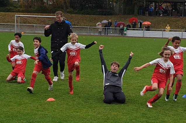 Gruppenszene von jubelnden Kindern in roten Fu&szlig;balltrikots auf einem Rasenplatz, einige Spieler liegen und anderen feiern.