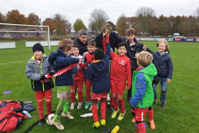 Gruppe von Kindern in Fu&szlig;balloutfits, die gemeinsam einen Pokal auf einem Sportplatz feiern.