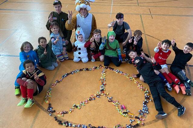 Gruppe von Kindern und Jugendlichen posiert um ein aus S&uuml;&szlig;igkeiten gelegtens Friedenssymbol in einer Sporthalle.