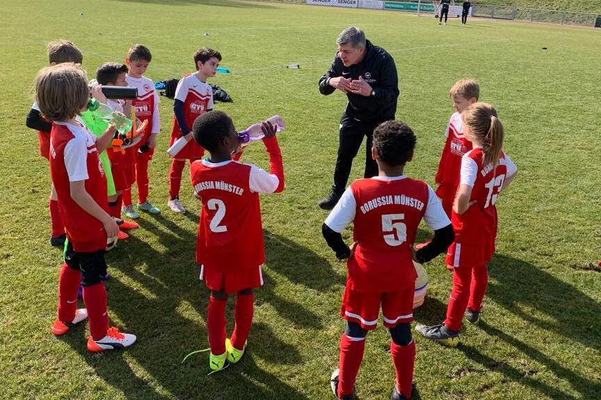 Trainer spricht mit einer Gruppe junger Fu&szlig;ballspieler in roten Trikots auf einem Rasenfeld.