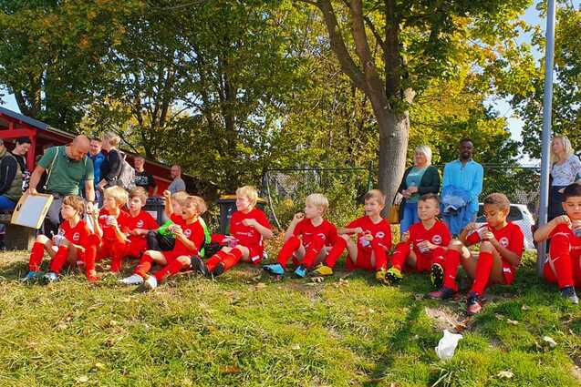 Gruppe von Kindern in roten Fu&szlig;balltrikots sitzt auf einer Wiese, Zuschauer im Hintergrund. Stra&szlig;e im Bild beschildert.