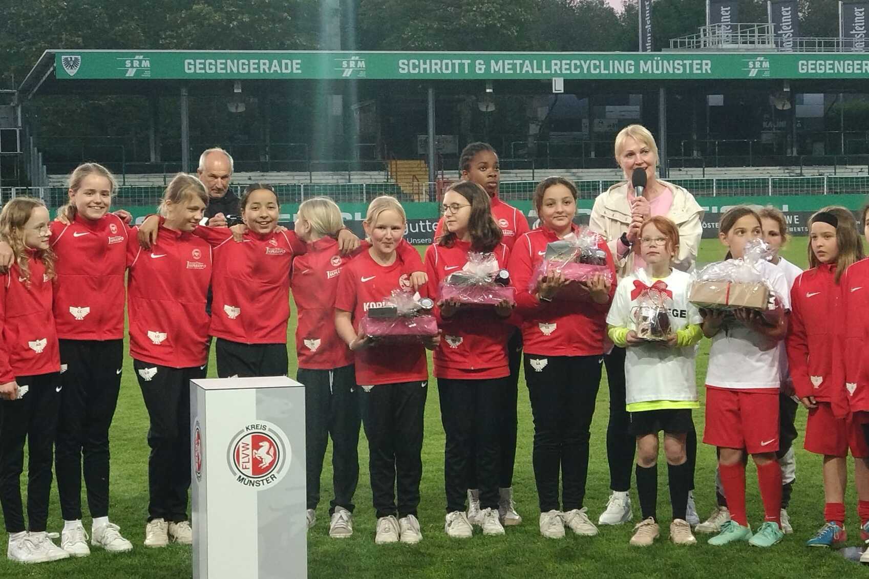 Mädchenmannschaft in roten Trainingsanzügen sammelt sich auf dem Spielfeld für eine Ehrung mit Präsentkörben.