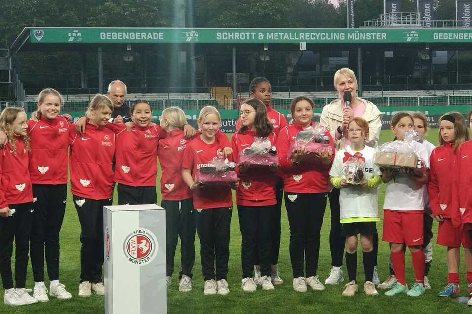 M&auml;dchenmannschaft in roten Trainingsanz&uuml;gen sammelt sich auf dem Spielfeld f&uuml;r eine Ehrung mit Pr&auml;sentk&ouml;rben.