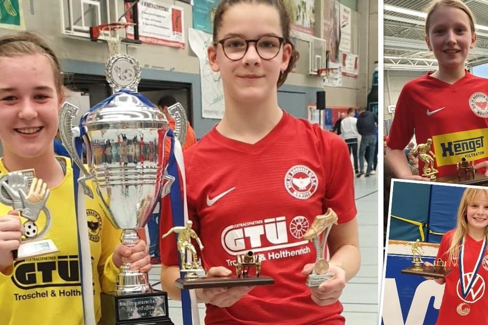 Drei junge M&auml;dchen in Sportkleidung halten Pokale und Medaillen nach einem Fu&szlig;ballturnier in einer Halle.