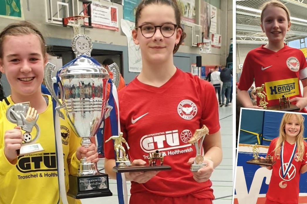 Drei junge M&auml;dchen in Sportkleidung halten Pokale und Medaillen nach einem Fu&szlig;ballturnier in einer Halle.